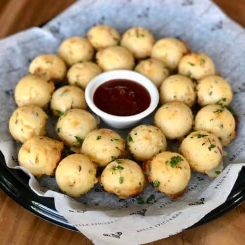 A platter of bite-sized cheesy bread balls, garnished with herbs, served with a side of dipping sauce.