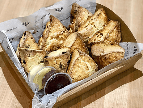 A basket of freshly baked scones, served with small containers of jam and cream, lined with parchment paper.