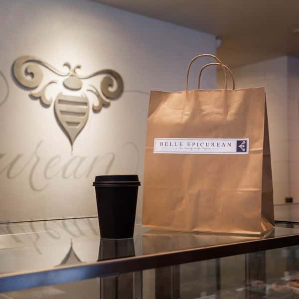 A brown paper bag with a "Belle Epicurean" logo on it, accompanied by a black coffee cup, set against a store wall with the brand's bee logo.