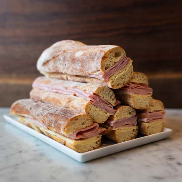 A stack of fresh sandwiches made with ciabatta bread and filled with layers of sliced ham, served on a white plate.