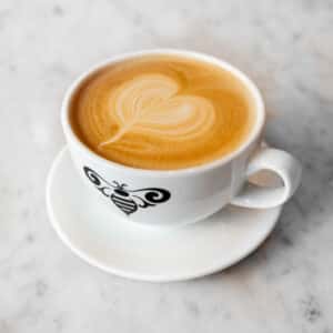 A cup of latte with a heart-shaped foam design, featuring a bee logo on the side, served on a white saucer.