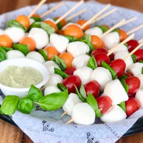 A platter of skewers with mozzarella balls, cherry tomatoes, and fresh basil, served with a side of dipping sauce.