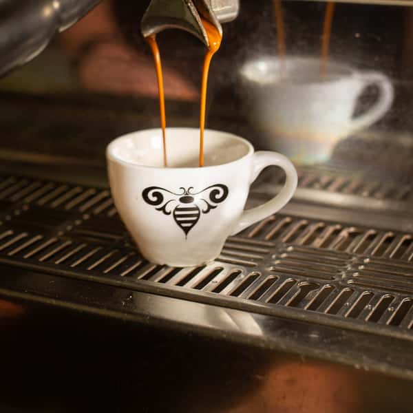 Espresso being poured from a coffee machine into a white cup with a bee logo, with the machine's reflective surface in the background.