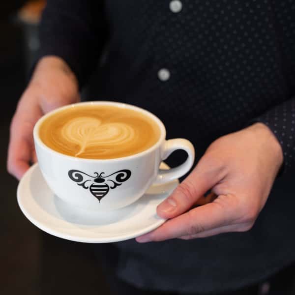 A person holding a cup of latte with a heart-shaped foam design, featuring a bee logo on the side, served on a white saucer.