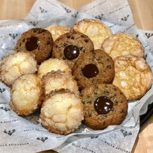 A selection of freshly baked cookies, including chocolate chip, coconut, and sugar-coated varieties, arranged in a basket with parchment paper.