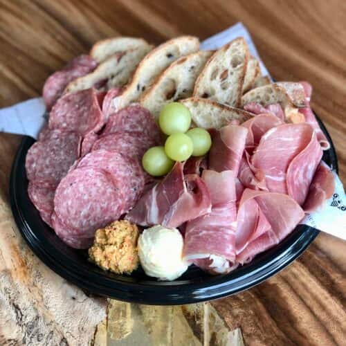 A platter of sliced cured meats, including salami and prosciutto, served with bread slices, grapes, and a side of mustard or dip.