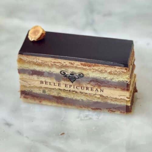 A layered cake with chocolate and cream fillings, topped with a whole hazelnut, and featuring a branded logo.