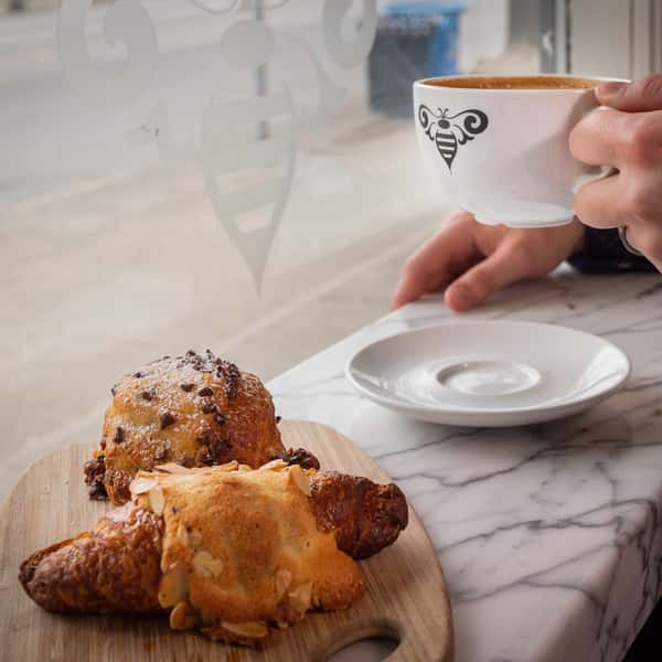 A person holding a cup of coffee with a bee logo, sitting next to a wooden board with a croissant and pastry. The scene is seen through a window.