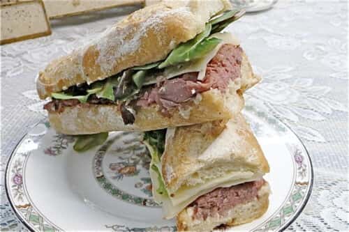 A close-up of two sandwiches stacked on a plate, filled with slices of roast beef, cheese, and fresh greens, served on crusty baguette-style bread.