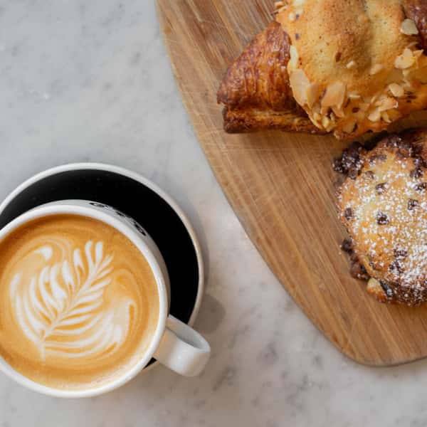 A cup of latte with leaf-shaped foam art, accompanied by a croissant and a pastry on a wooden board.