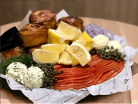 A platter featuring smoked salmon, lemon wedges, capers, herbed butter, and freshly baked pastries arranged on parchment paper.