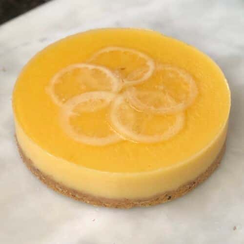 A round lemon cheesecake with a smooth yellow topping, decorated with thin slices of lemon on top.