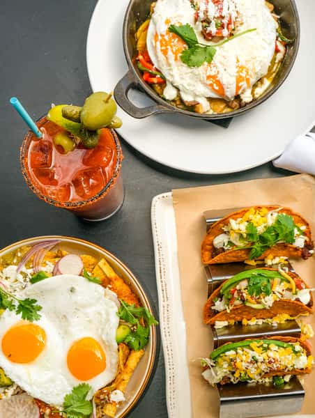 Mexican brunch spread featuring street tacos, chilaquiles topped with fried eggs, and a spicy Bloody Mary cocktail.