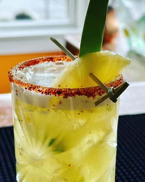 Pineapple cocktail with chili rim, garnished with a pineapple wedge and leaf in a tall glass filled with ice.
