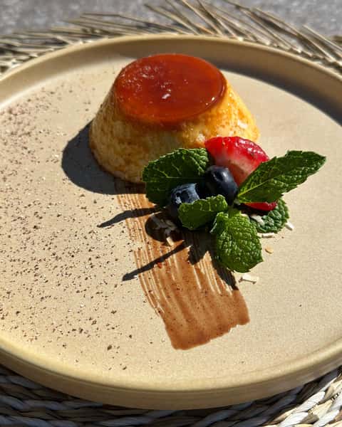 Caramel flan served with fresh berries, mint leaves, and a chocolate smear on a beige plate.
