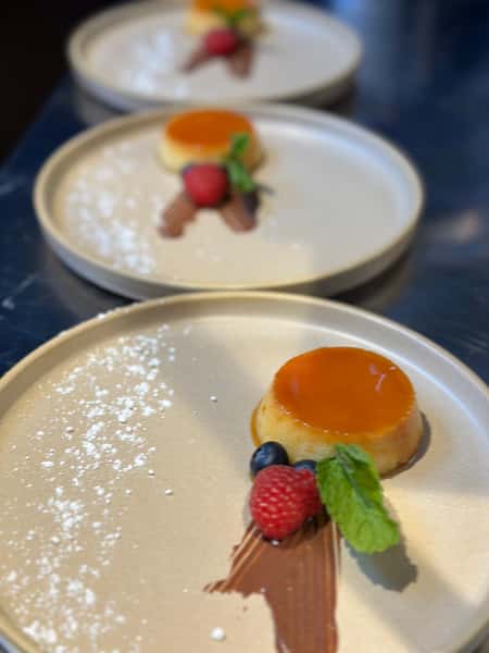 Creamy caramel flan served with a swipe of chocolate sauce, fresh berries, and mint on a beige plate dusted with powdered sugar.