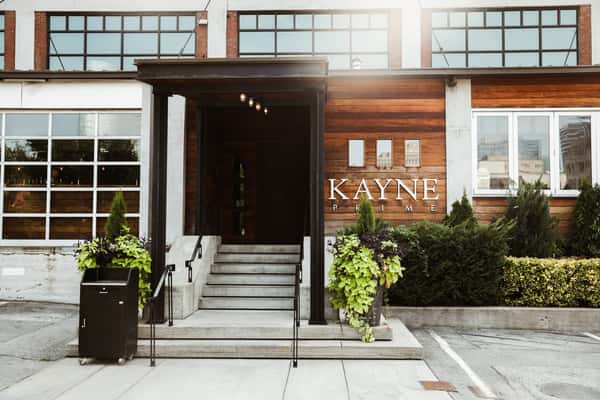 Facade of Kayne Prime, an upscale American Steakhouse, with wooden panels, bold name signage, and welcoming steps framed by lush greenery, reflecting its modern yet classic dining ambiance.