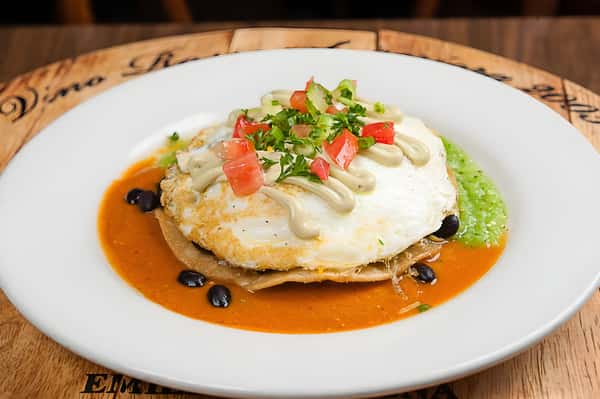 Huevos Rancheros or a similar egg dish, featuring a fried egg over a tortilla with black beans, salsa, and a side of green avocado crema.