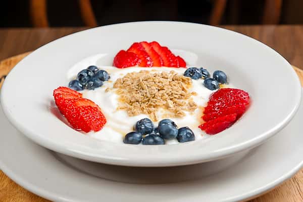 A yogurt parfait bowl featuring a central scoop of granola surrounded by fresh strawberries and blueberries.