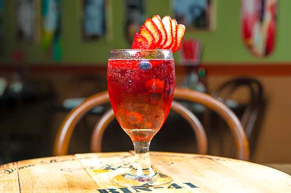 A vibrant red cocktail (possibly a sangria or fruit punch) garnished with thin apple slices, served in a stemmed glass on a wooden table.