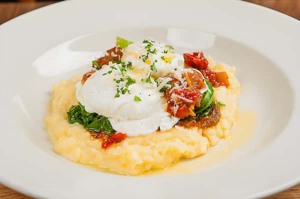 A poached egg dish served over a bed of yellow polenta or grits, garnished with sautéed greens and sun-dried tomatoes.