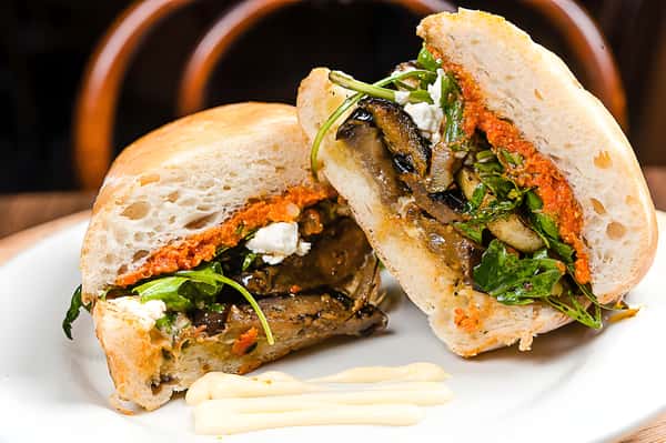 A gourmet sandwich on a ciabatta roll, sliced to reveal roasted vegetables (likely eggplant and peppers) and a bright orange spread or pesto.