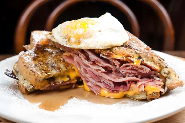 A hearty breakfast sandwich or Monte Cristo, sliced in half to show layers of ham and melted cheese, topped with a sunny-side-up egg.