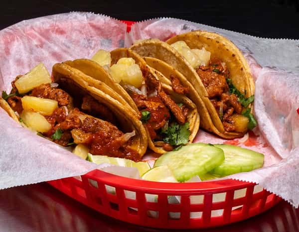 Four tacos al pastor filled with marinated pork, pineapple chunks, onions, and cilantro in soft corn tortillas, served in a red basket with cucumber slices and lime wedges.