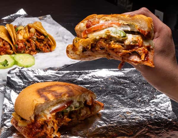Grilled torta al pastor with melted cheese, onions, jalapeños, and tomatoes held in hand, with a second sandwich and tacos al pastor on foil in the background.
