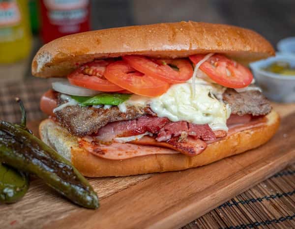 Juicy beef torta with melted cheese, ham, crispy bacon, fresh tomato, onions, and avocado on a soft telera roll, served with roasted jalapeños and salsa cups.