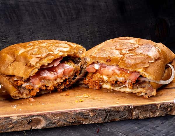 Grilled Mexican pambazo sandwich cut in half, stuffed with spicy chorizo, potatoes, melted cheese, tomato slices, and onions on a crispy guajillo chili-soaked roll — a bold and flavorful street food favorite from central Mexico.