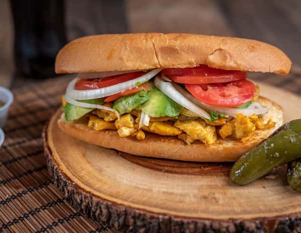 Mexican chicken torta sandwich filled with grilled chicken, sliced avocado, tomatoes, onions, and jalapeños on a toasted bolillo roll, served on a rustic wooden board with pickled peppers.