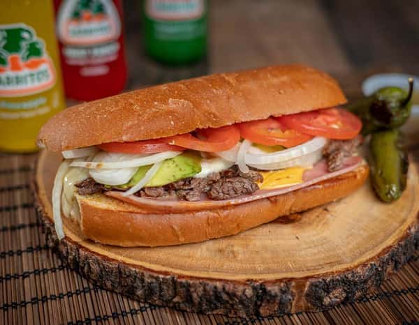 Traditional Mexican torta with thinly sliced steak, ham, avocado, tomato, onion, and cheese, served on a rustic wood slab with a grilled jalapeño.