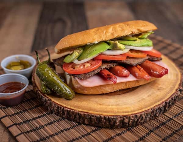Loaded Mexican hot dog torta packed with sliced hot dogs, steak, avocado, tomato, onion, and ham, served on a crusty roll with grilled jalapeños and salsas.