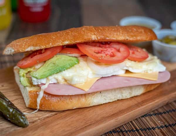 Mexican torta sandwich with sliced ham, American cheese, fresh avocado, grilled white cheese, onions, and ripe tomatoes on a toasted bolillo roll, served with a roasted jalapeño and salsas on the side.