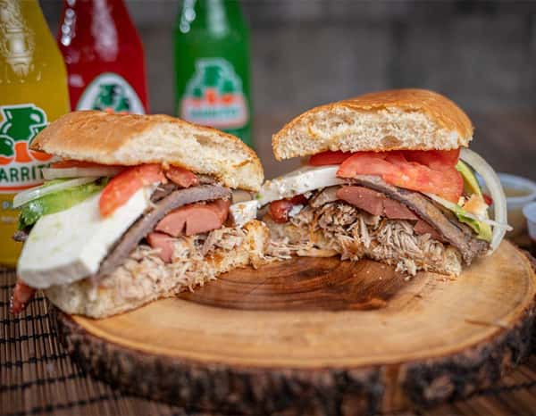 Split Mexican torta piled with shredded pork, grilled beef, hot dog slices, tomato, onion, avocado, and cheese on a fluffy bolillo roll, served on a wooden slab with colorful Jarritos sodas in the background — a bold, street-style sandwich perfect for authentic Mexican eateries.