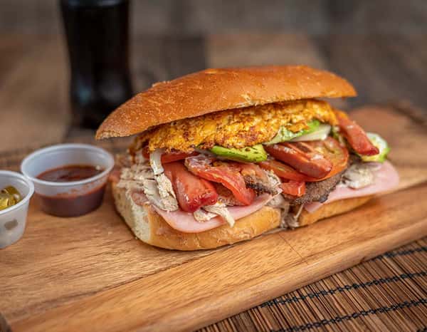 Mexican torta filled with ham, shredded pork, hot dog slices, grilled beef, avocado, tomatoes, onions, and an egg omelet, served on a toasted bolillo roll with salsa and jalapeños on the side.