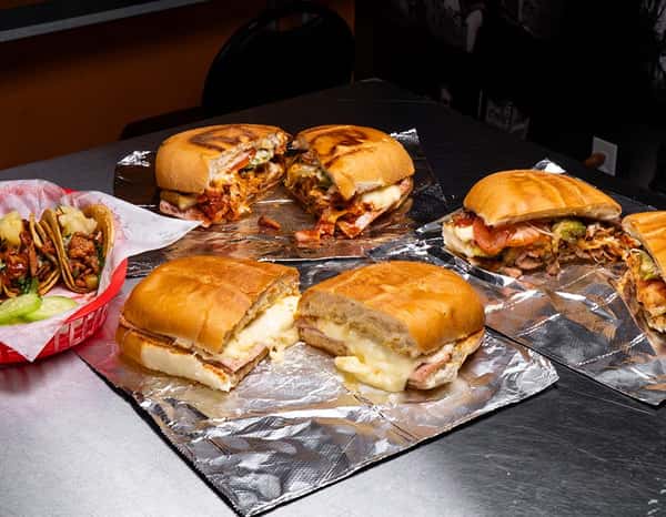 Assorted Mexican tortas on foil-lined trays, showcasing variations with cheese, ham, al pastor, and sausage, next to a basket of tacos with pineapple and cucumber slices.