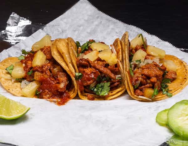 Three tacos al pastor filled with marinated pork, pineapple chunks, onions, and fresh cilantro on corn tortillas, served on parchment with lime and cucumber slices — a classic Mexican taco plate bursting with flavor.