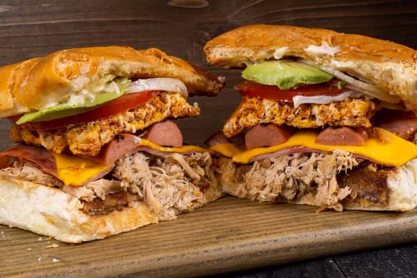 Hearty Mexican torta cut in half, stacked with pulled pork, grilled hot dog slices, cheddar cheese, ham, spiced egg patty, avocado, tomato, onions, and a smear of mayonnaise on toasted telera bread — a bold and flavorful sandwich perfect for Mexican street food lovers and torta shop menus.