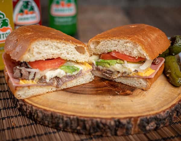 Sliced torta mexicana with ham, grilled beef, cheese, avocado, tomatoes, and onions, served on a rustic wooden board with pickled jalapeños and colorful Jarritos sodas in the background.