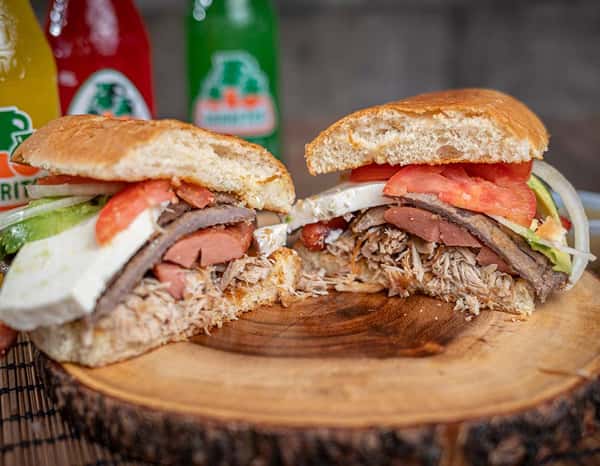 Cross-section of a hearty Mexican torta stuffed with shredded pork, steak, avocado, tomato, onion, hot dog slices, and panela cheese, served on a wood-cut board with Jarritos soda in the background.