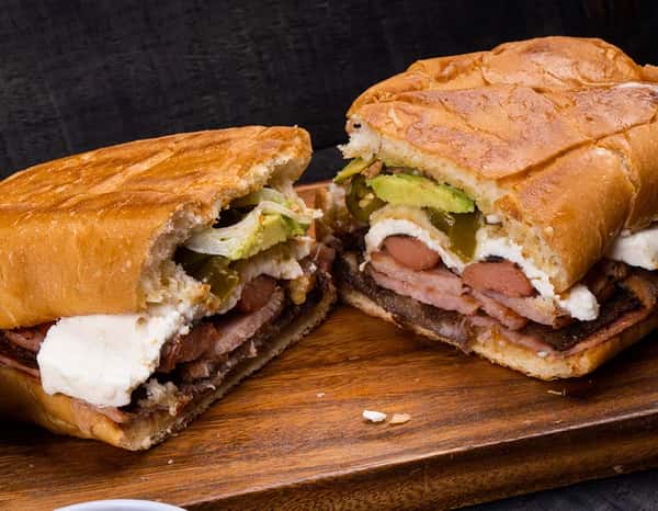 Hearty Mexican torta sandwich split in half, loaded with grilled meats, sliced avocado, jalapeños, creamy cheese, and fresh lettuce on toasted bolillo bread.