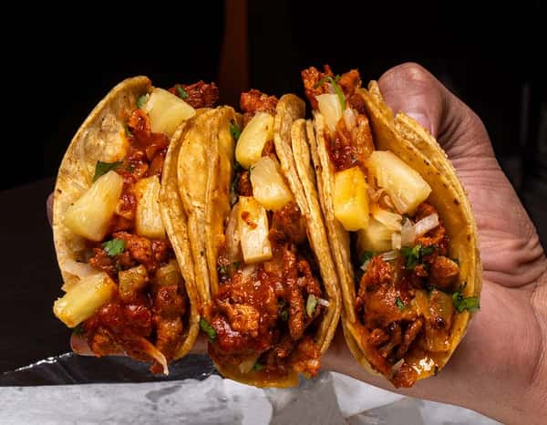 Hand holding three tacos al pastor filled with marinated pork, grilled pineapple chunks, chopped onions, and fresh cilantro on warm corn tortillas — a classic Mexican street food bursting with smoky, sweet, and savory flavors.