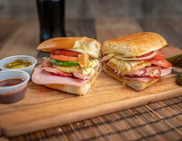 Split Mexican torta showcasing layers of grilled ham, pineapple, sliced tomato, onions, avocado, and melted cheese on a toasted bun, with salsa and jalapeños on the side.