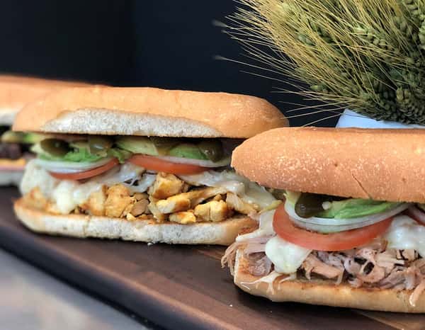 Two Mexican torta sandwiches filled with grilled chicken, shredded pork, melted cheese, fresh avocado, tomato, jalapeños, and onions, served on crusty bolillo rolls.