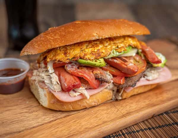 Overloaded Mexican torta with grilled meats, ham, hot dogs, eggs, tomato, avocado, and melted cheese on a toasted bolillo roll with a side of salsa.