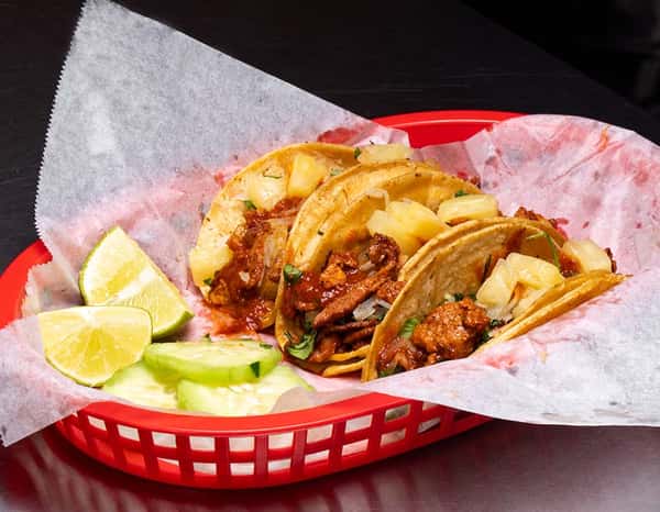 Basket of tacos al pastor topped with juicy pork, fresh pineapple chunks, onions, and cilantro on soft corn tortillas, garnished with lime wedges and cucumber slices.