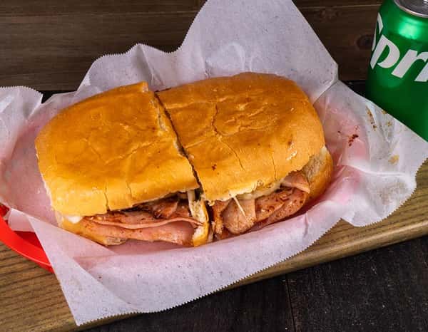 Classic Mexican torta sandwich wrapped in deli paper and served in a red basket, stuffed with grilled ham, sausage, and melted cheese on a toasted bolillo roll, with a can of Sprite in the background.