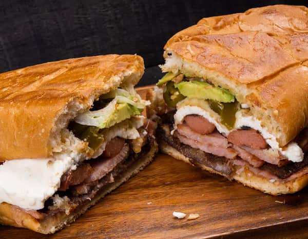 Juicy Mexican torta sliced in half, showcasing layers of grilled beef, ham, hot dogs, fresh avocado, jalapeños, and creamy cheese on crispy bread.